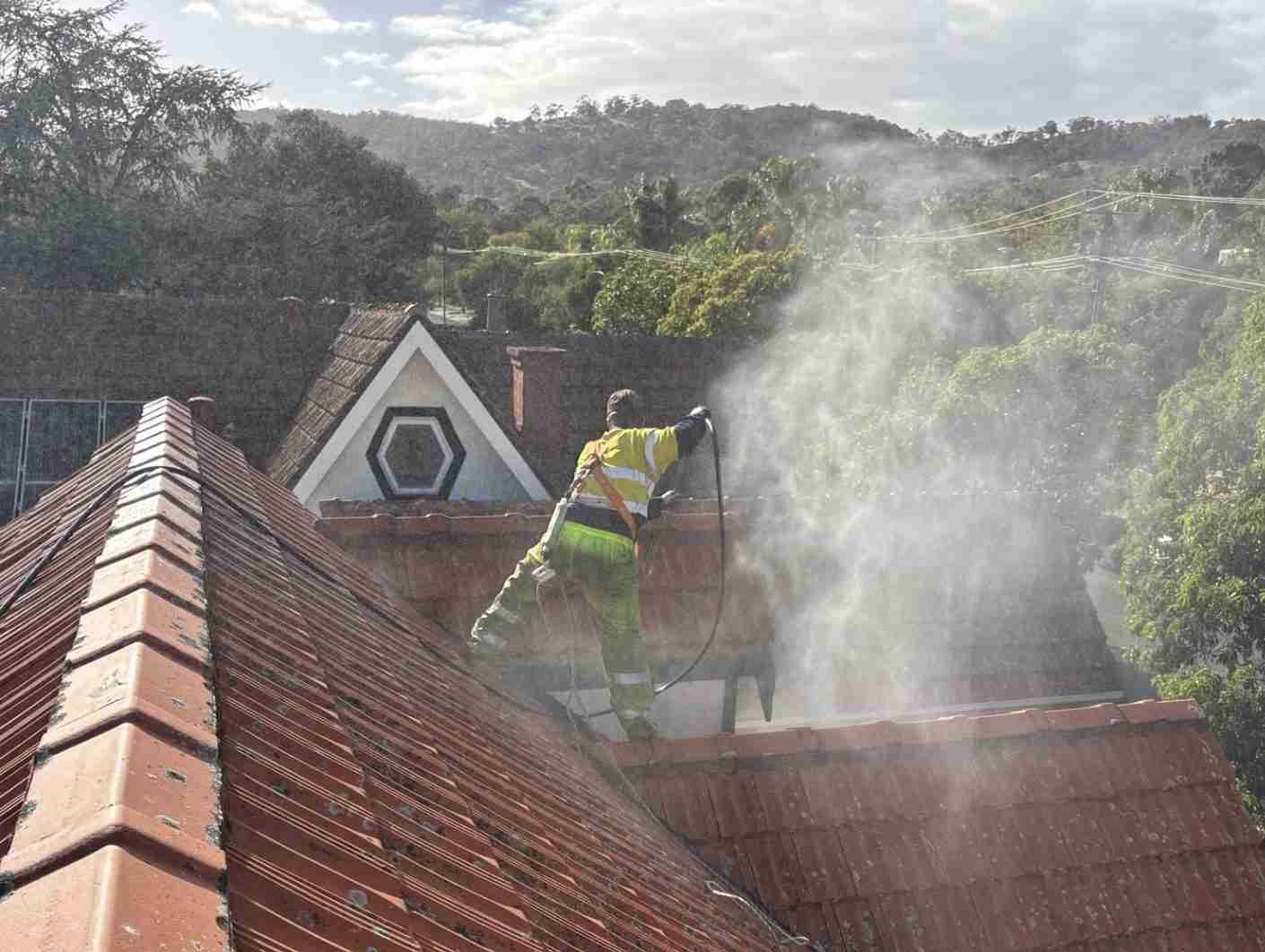 A person is standing on a roof in a harness pressure-cleaning the roof