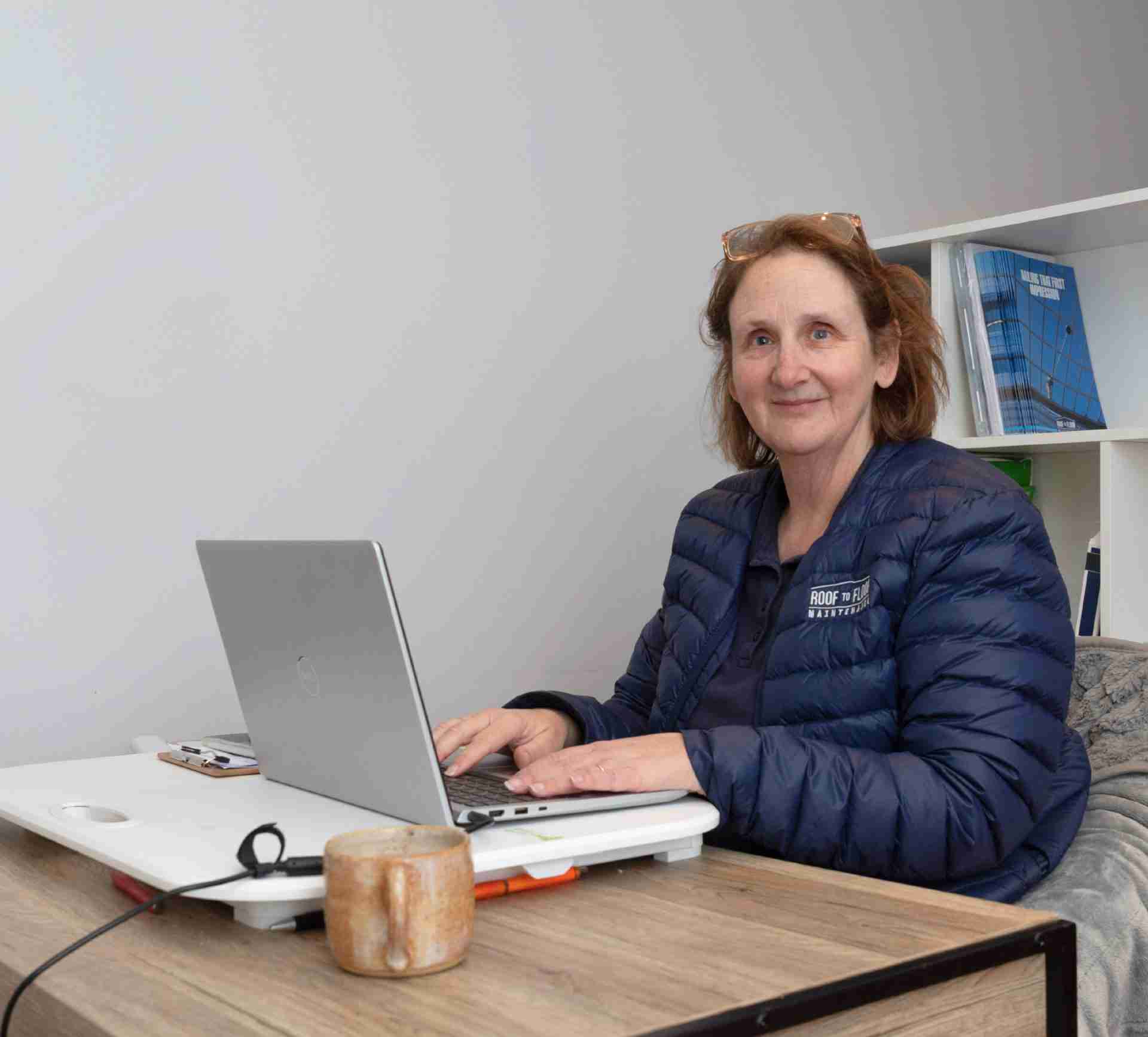 A woman sits behind a laptop with her hands typing on the keyboard. She is smiling and wearing a blue Roof to Floor Maintenance puffer jacket
